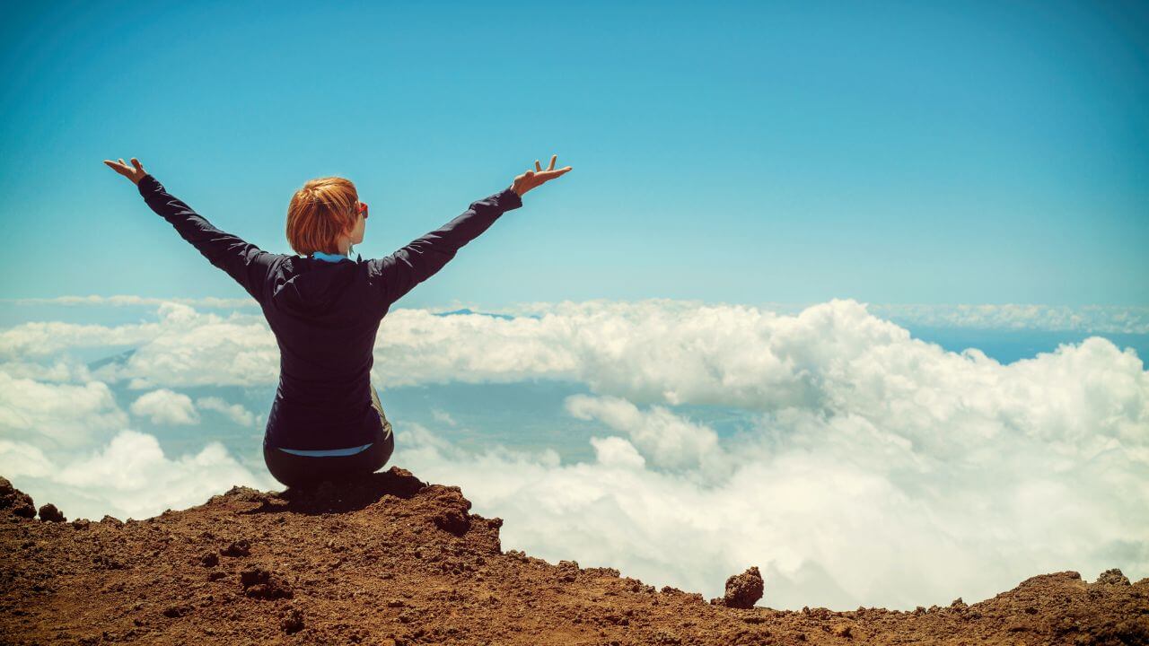 mulher de braços abertos, sentada no alto de uma montanha que está acima das nuvens. Ela demonstra liberdade, saúde mental e bem estar